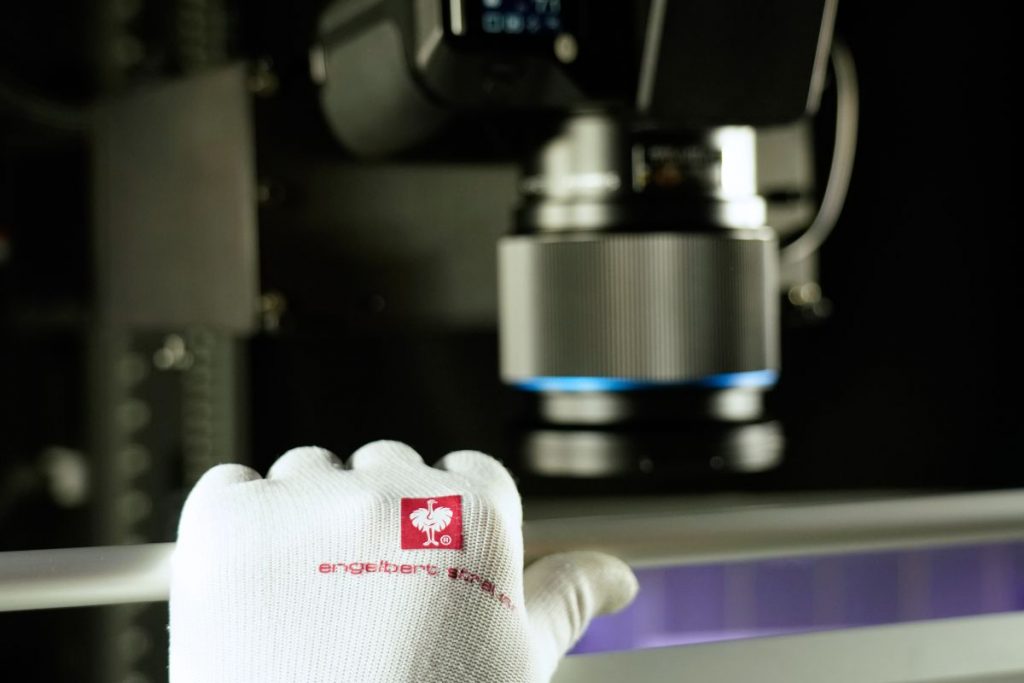 Eine Hand mit weißem Habdschut senkt eine Glasplatte. Im Hintergrund ist eine hochwertige Reproeinrichtung mit Kamera zu sehen.